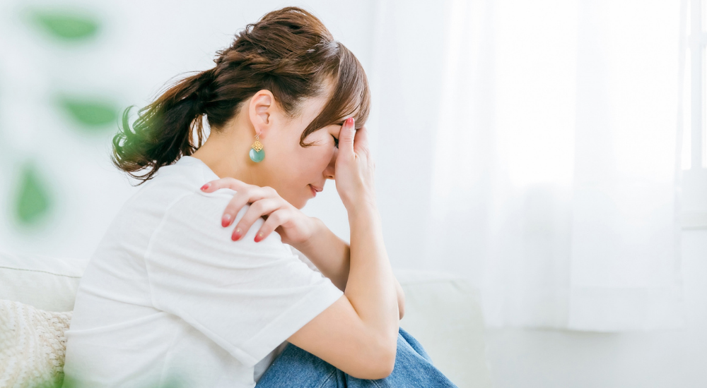 ソファに座り、【首から肩（僧帽筋）】にかけてを手で押さえ、慢性的な痛みやこりで辛そうにしている女性。