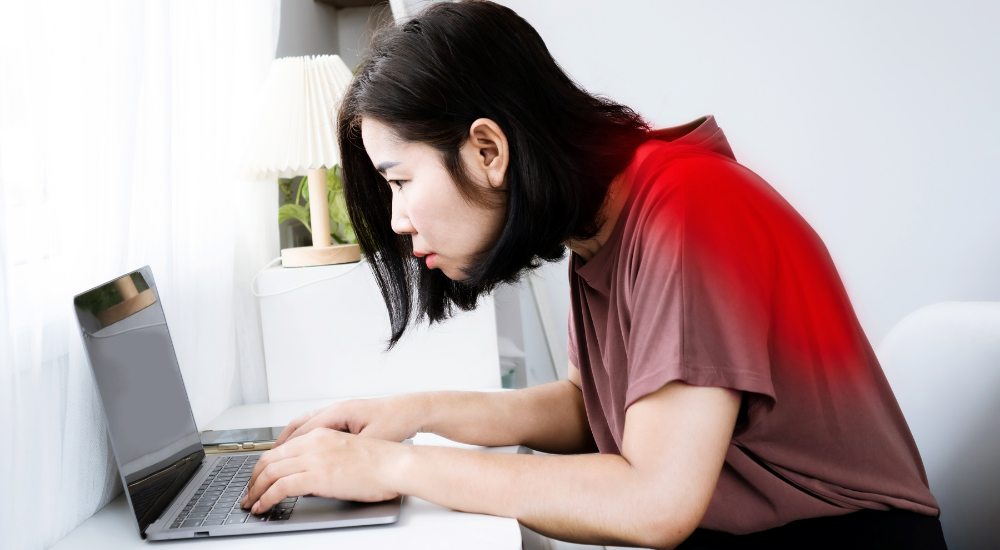 デスクワーク中に首を前に突き出した女性の背中が赤く発光している画像で、【頸部・背部（僧帽筋・肩甲挙筋・板状筋周辺）】における長時間の不良姿勢（ストレートネックや猫背）が引き起こす、ガンコな首こりや背中の強い張り、痛みの原因である猫背姿勢、ストレートネックを解説している様子。