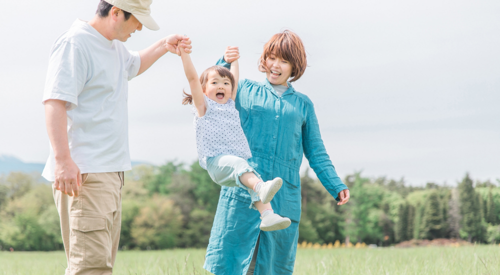 緑豊かな公園で両親が幼い娘の両手を引いて元気に持ち上げて遊んでいる画像で、【腰部・肩関節（脊柱・ローテーターカフ周辺）】を示し、ぎっくり腰や肩の痛みの不安なく、安心して子供との触れ合いや育児を楽しめている健やかな日常の様子。