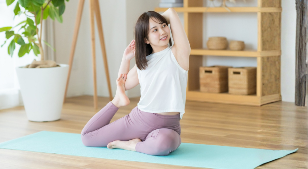 明るい部屋でヨガマットに座り、脚を曲げて腕を後頭部に回して体側や前ももを伸ばすポーズをとる女性の画像で、【股関節・体幹（腸腰筋・大腿四頭筋・広背筋周辺）】を示し、ヨガやストレッチによって縮こまった筋膜が心地よく引き伸ばされ、全身の柔軟性と血流が改善している様子。