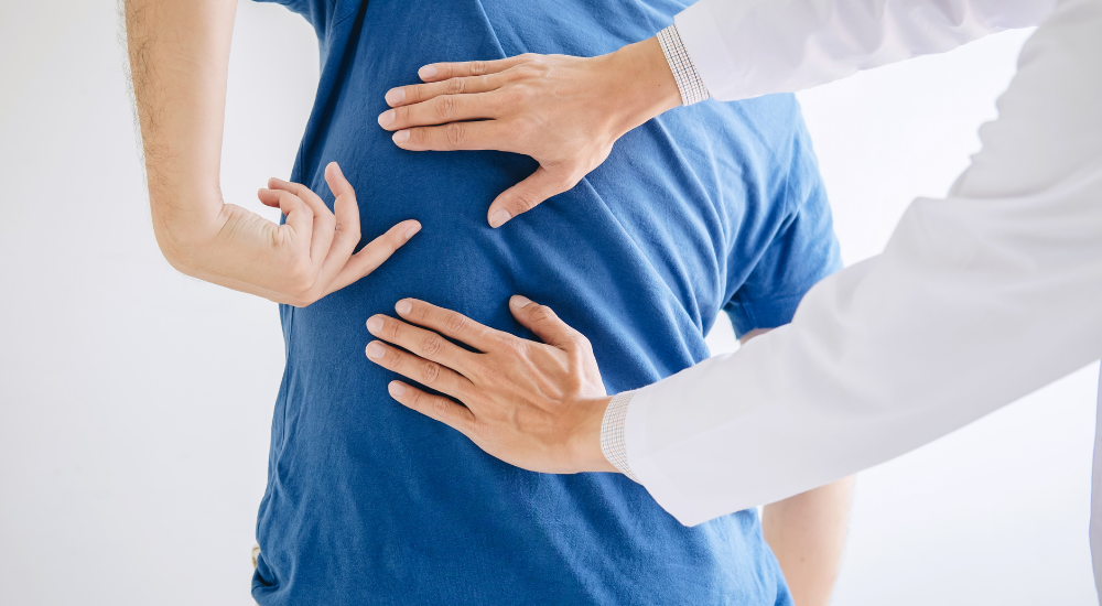 白衣を着た専門家（理学療法士・施術者）が、患者の【背中から腰（胸腰移行部・脊柱起立筋周辺）】に手を当てて、姿勢や筋肉の緊張状態を丁寧に触診・調整している様子。急性腰痛（ぎっくり腰）や急性背部痛（ぎっくり背中）を示した画像。