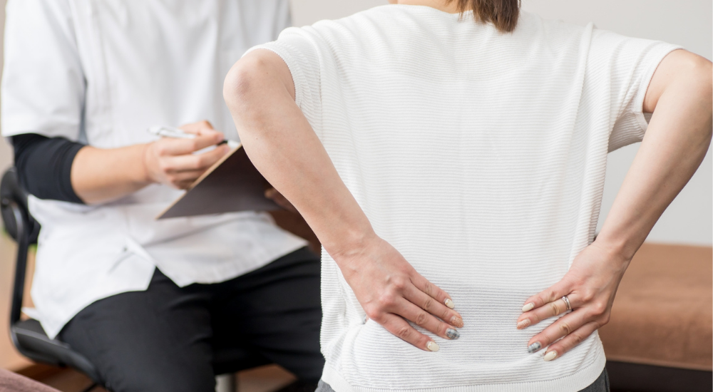 両手を腰に当て、【腰椎】や【仙腸関節】付近の痛みを専門家に説明している状況です。立ち上がりや日常動作で感じる腰への不安感や慢性的な辛さを、切実に打ち明けている風景です。